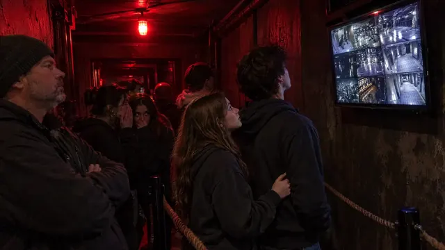 In the red-lit queue for a haunted attraction, a couple stare up at the monitor while friends behind them whisper