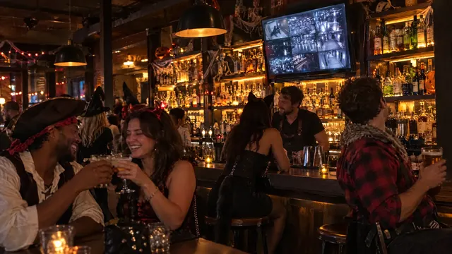 Friends in costume clink glasses at a busy bar while the monitor plays on a TV above the bottles